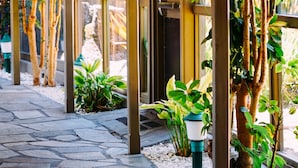 A covered walkway with stone flooring, surrounded by potted plants and trees.
