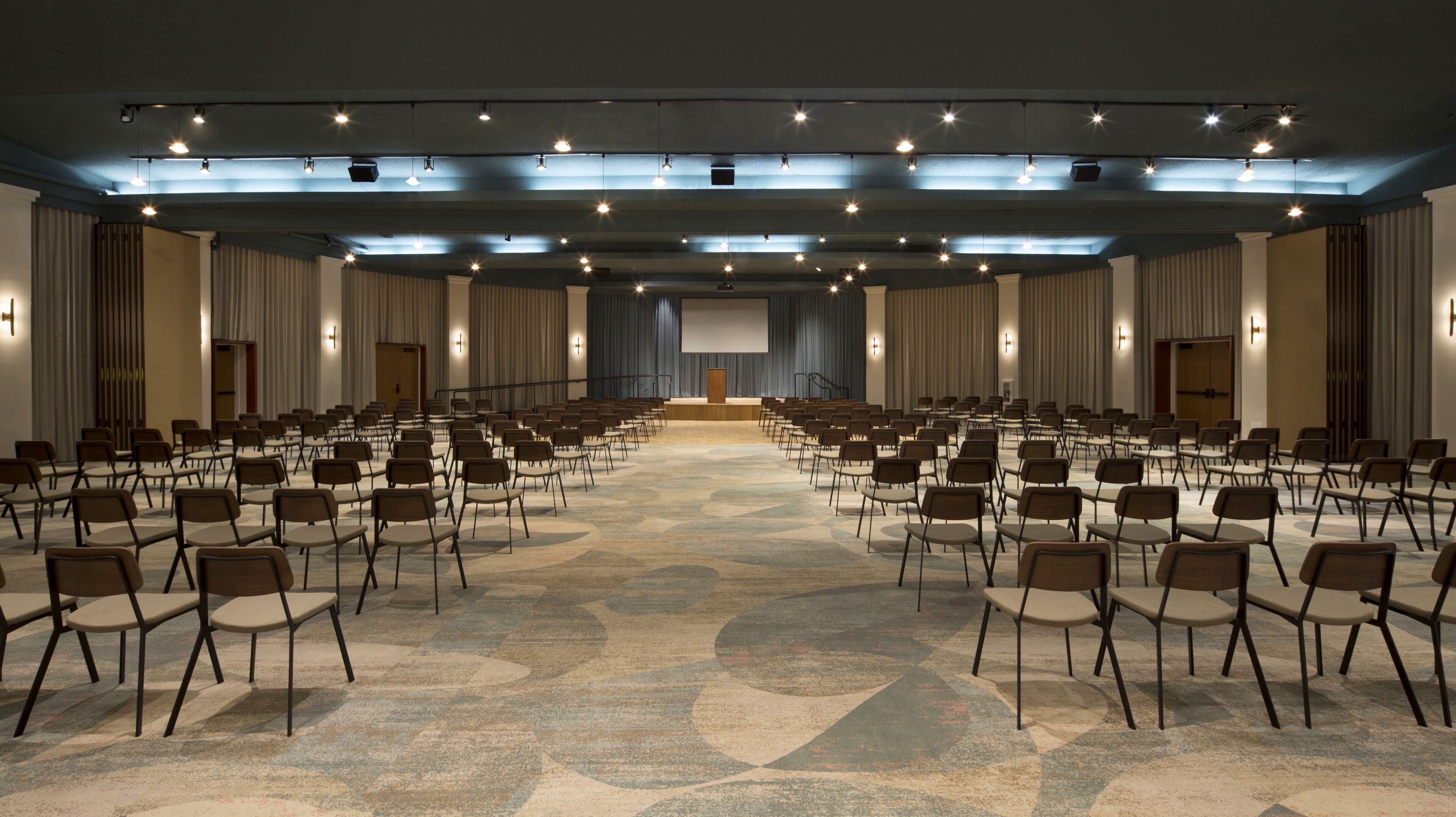 A large conference room with rows of chairs facing a stage, equipped with a projector and lighting.