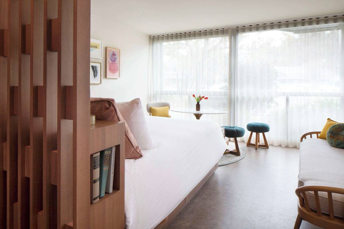 Habitación de hotel moderna con una cama grande, cabecera de madera, una mesita redonda con un florero y dos otomanas, con vistas a la vegetación a través de cortinas traslúcidas.