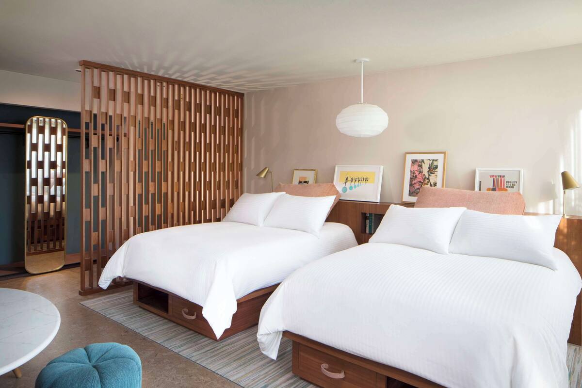 Habitación de hotel con dos camas, un separador de madera, una mesa redonda y una lámpara colgante.
