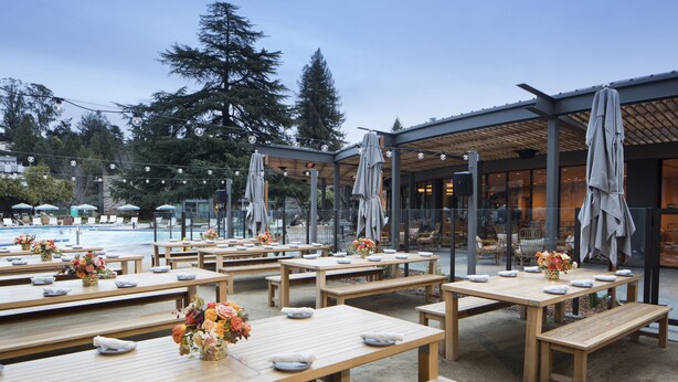 An outdoor dining area with wooden tables and benches, some with umbrellas, and a pool in the background.