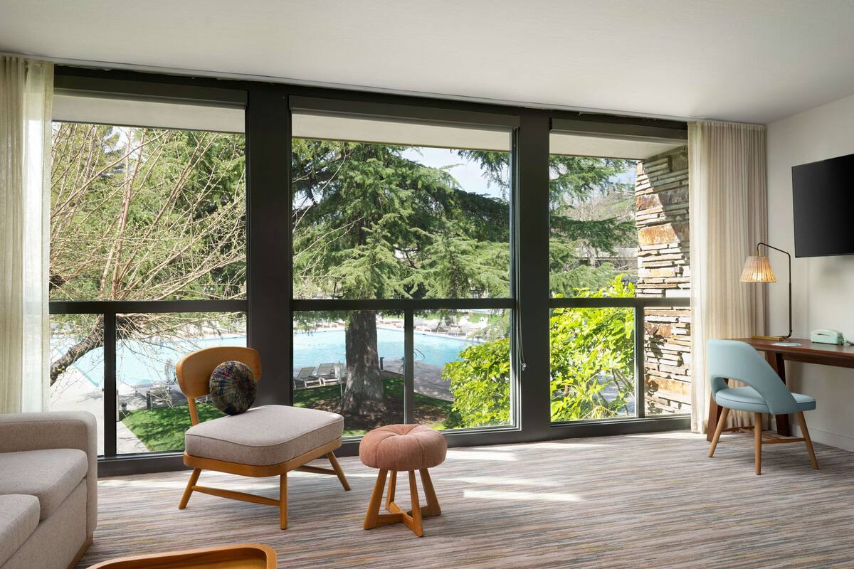 Una sala de estar moderna con un amplio ventanal corredera que da a una zona de piscina, un sofá, una silla, un pequeño reposapiés y un escritorio con una lámpara.