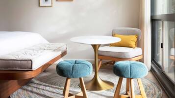 Una habitación de hotel moderna con una cama, una mesita pequeña y redonda, una silla y dos otomanas.