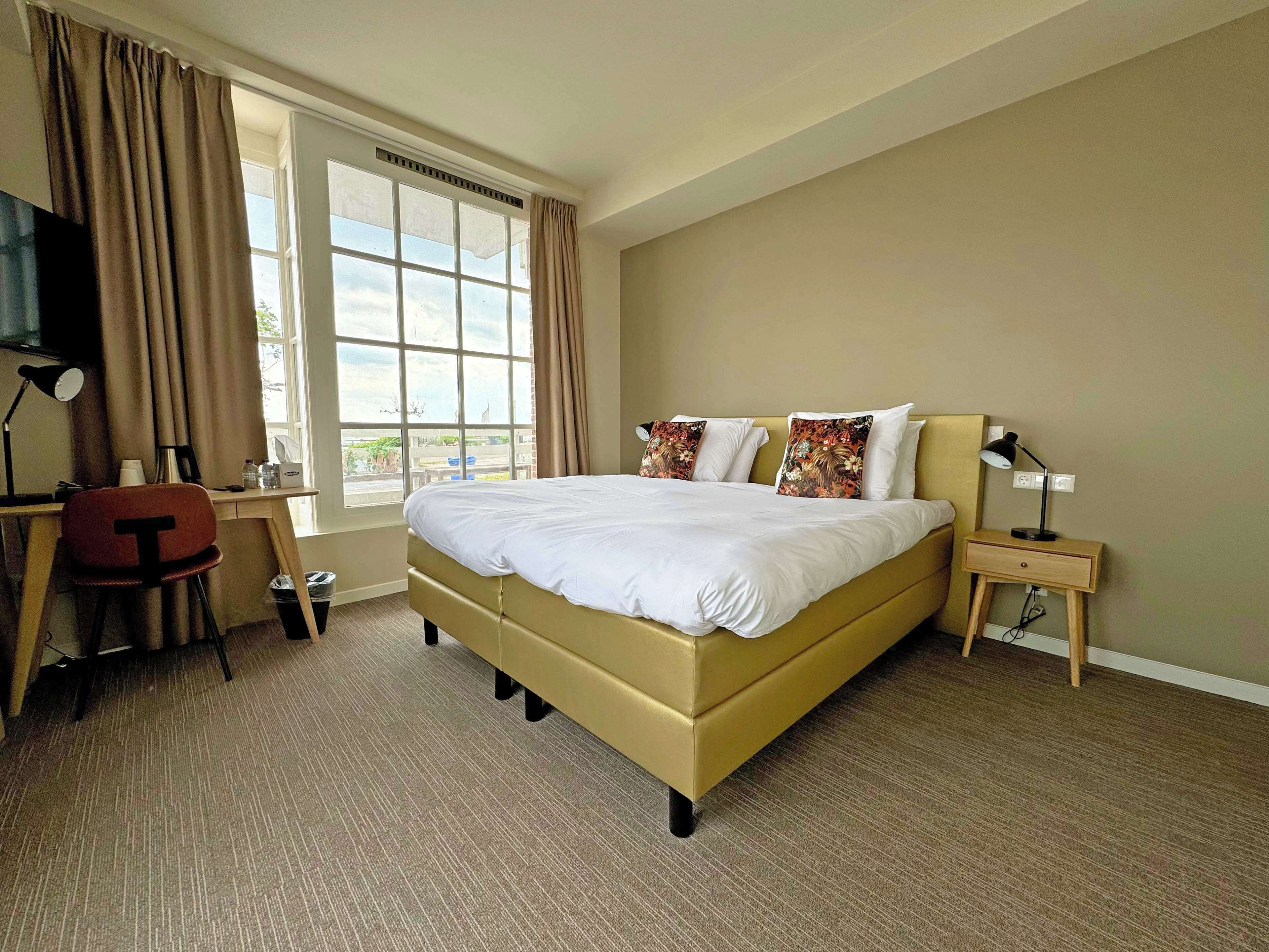 A hotel room with a large bed, a desk with a chair, a small bedside table, and a window with curtains.