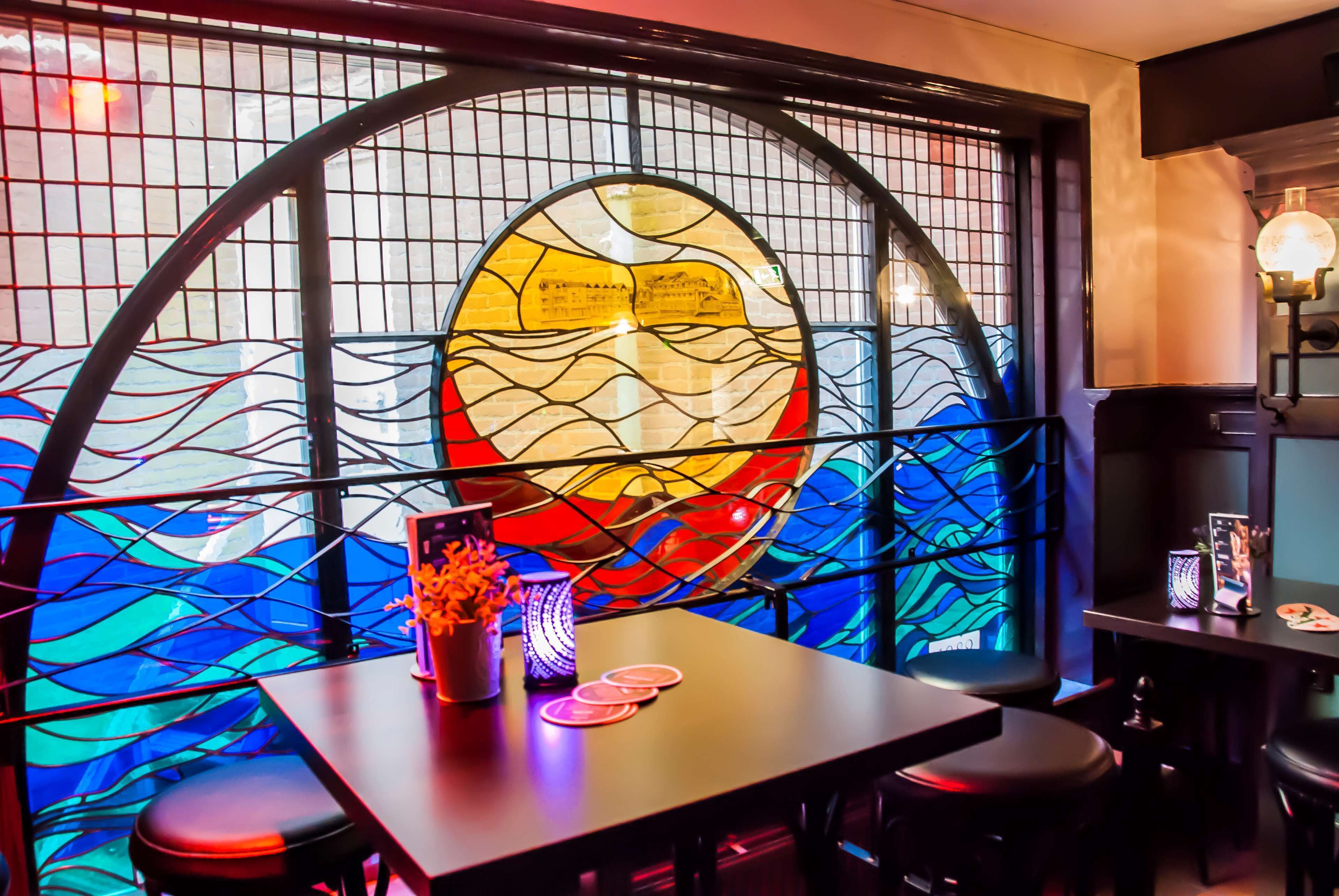 Un restaurante con un ventanal de vitral, una mesa con un jarrón y posavasos, y una zona de bar con taburetes y un televisor.