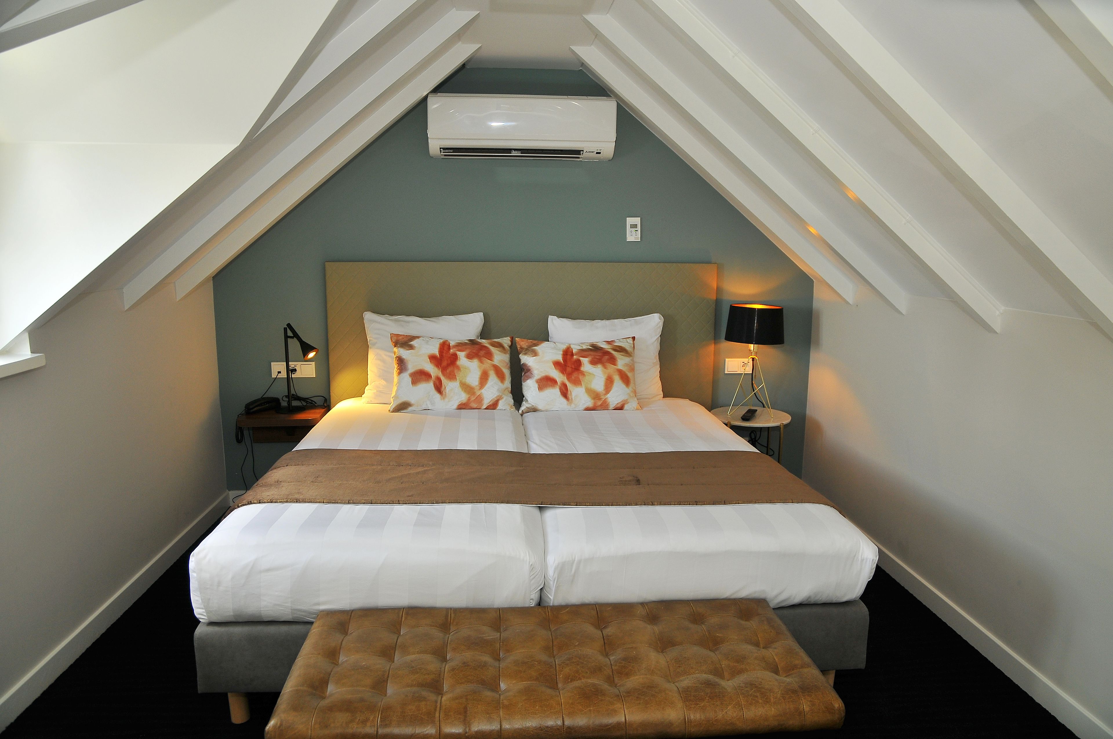Une chambre avec un plafond en pente, un lit avec une literie blanche et des coussins à motifs floraux, une table de chevet avec une lampe, et un banc au pied du lit.