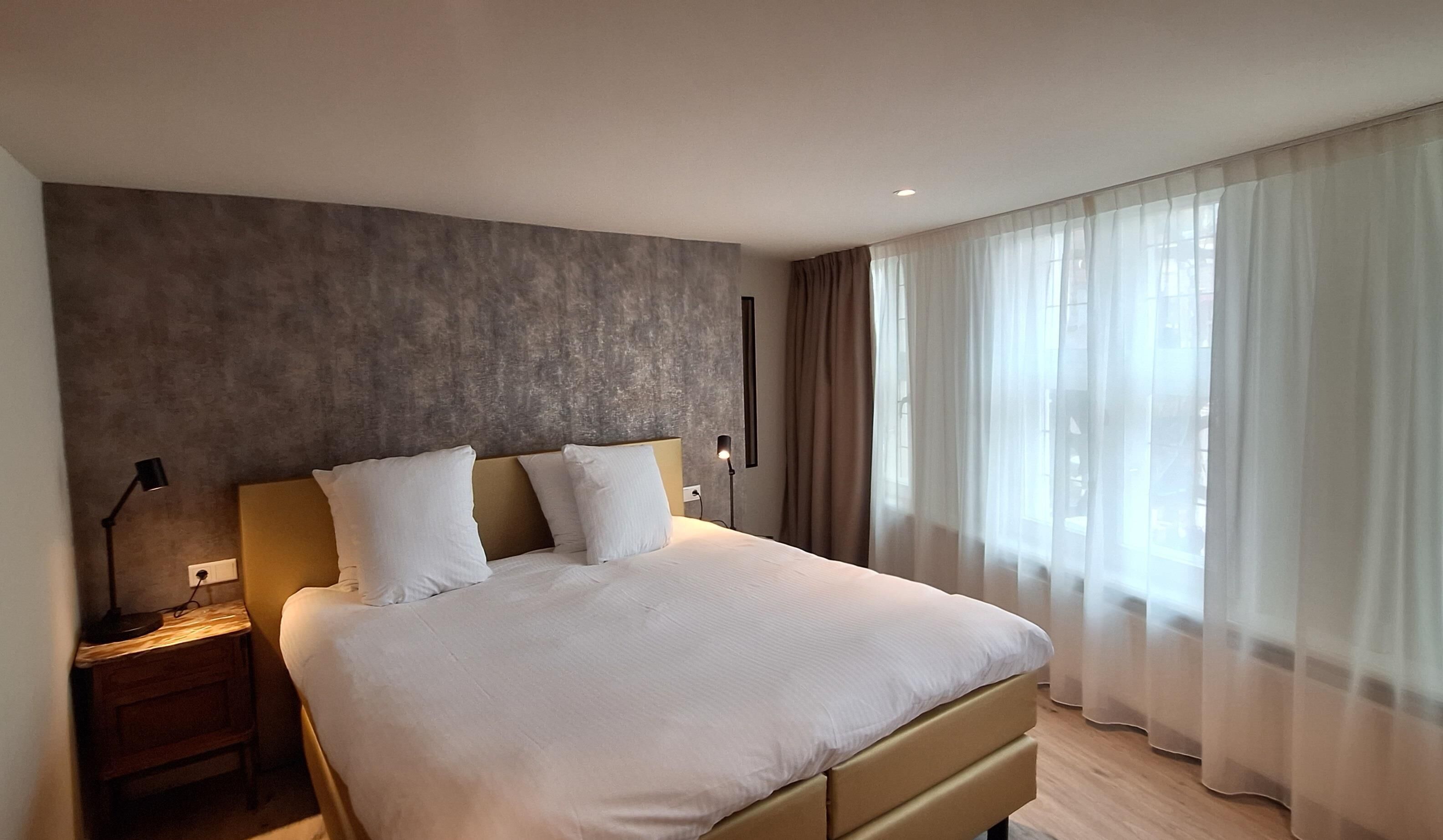 A modern hotel room with a large bed, a bedside table, a lamp, and sheer curtains.