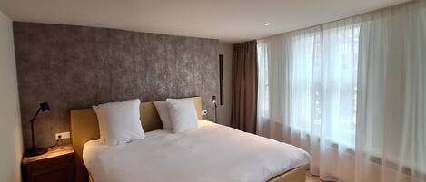 Una habitación de hotel moderna con una cama grande, una mesita de noche, una lámpara y cortinas traslúcidas.