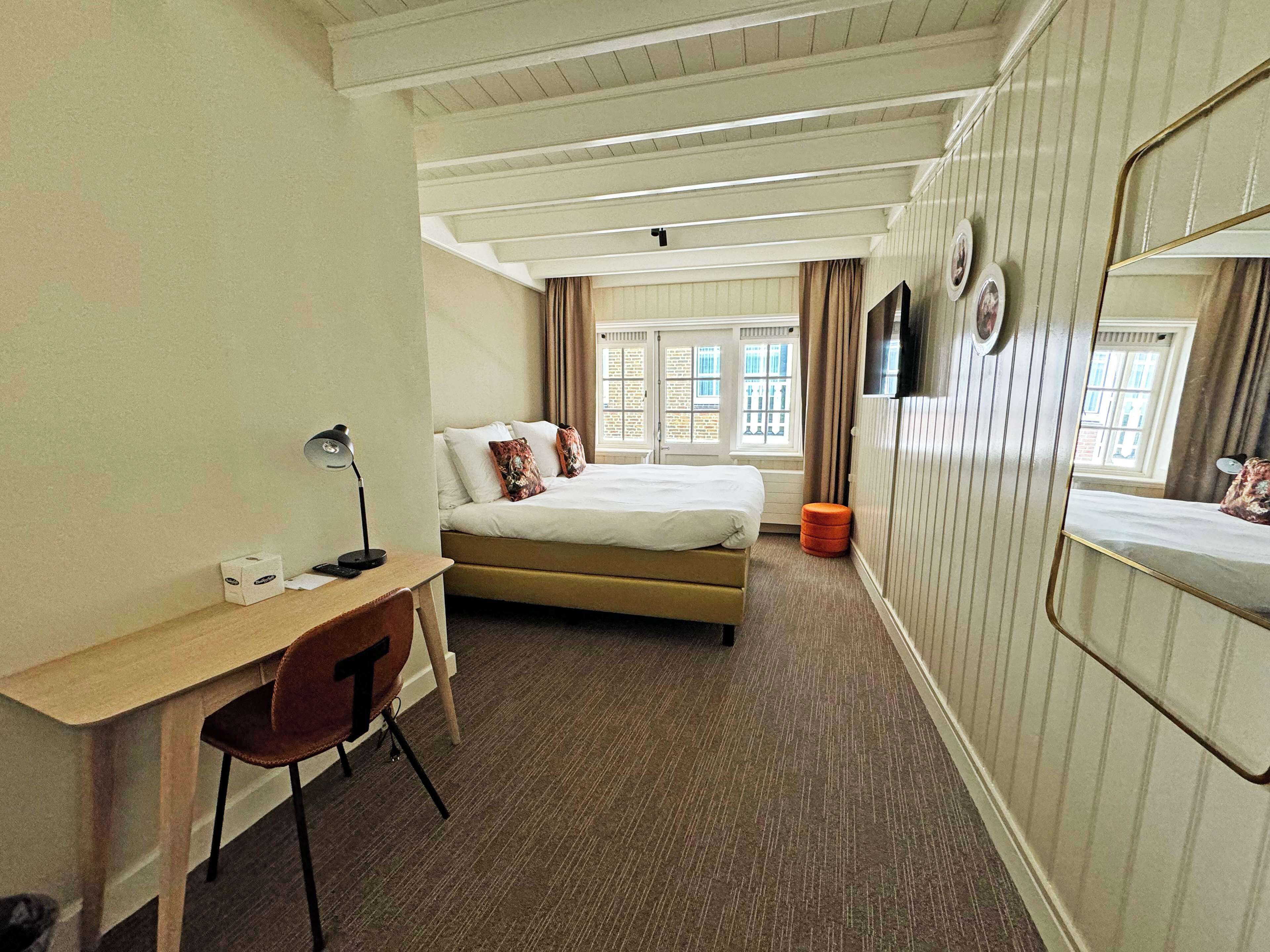 A hotel room with a bed, a desk, a chair, a mirror, and a window with curtains.