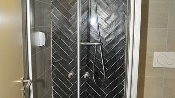 A walk-in shower with a glass door, black herringbone tile walls, and a white toilet in the adjacent bathroom.