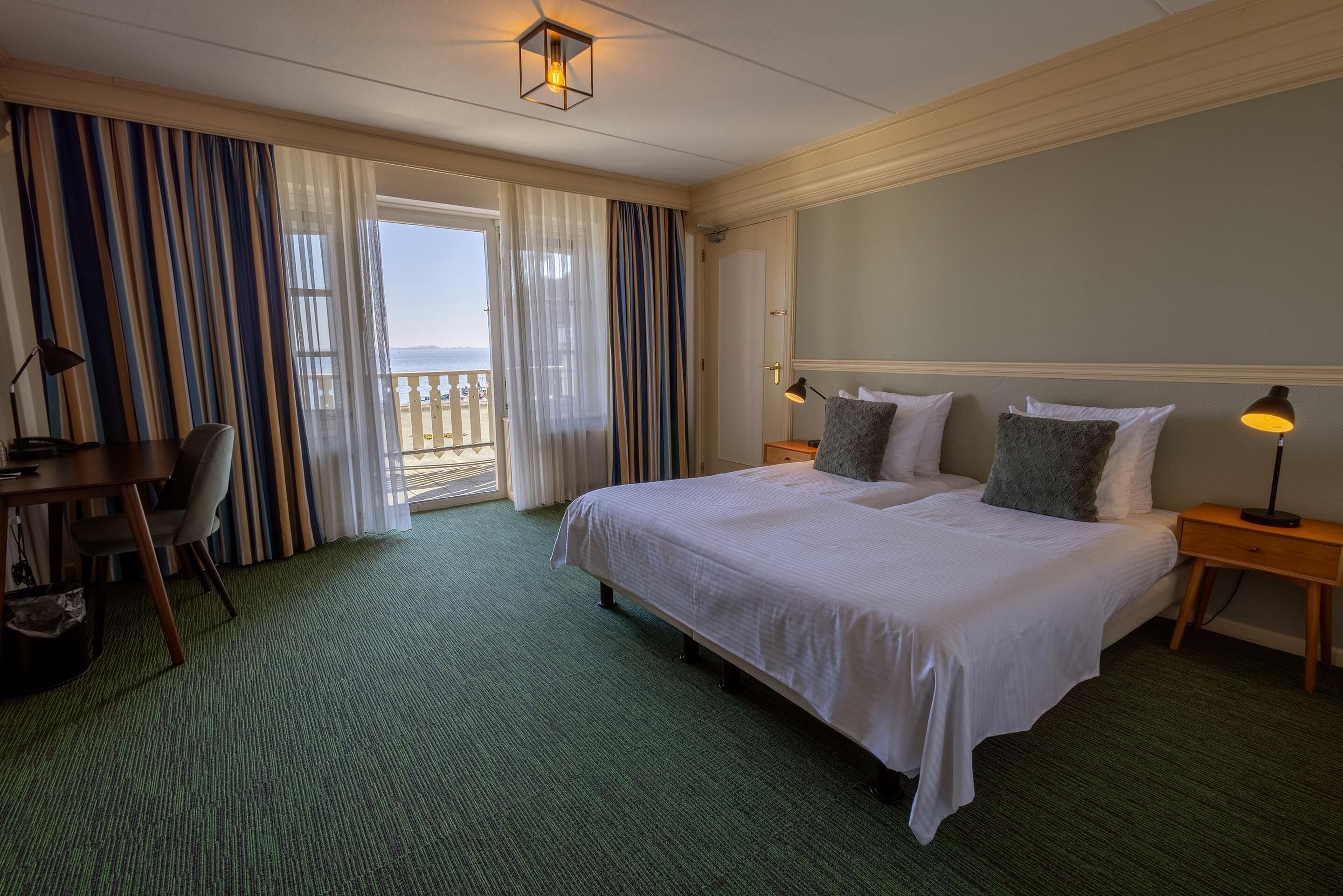 Habitación de hotel con una cama grande, un escritorio y una silla. Tiene un balcón con cortinas.