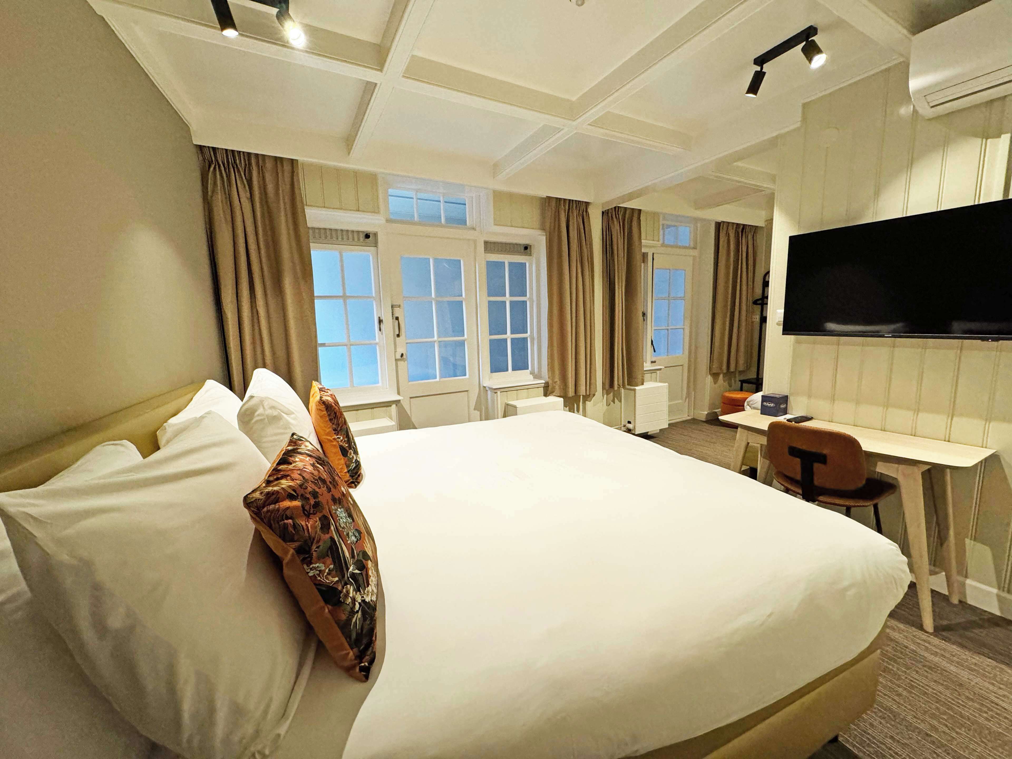 A hotel room with a large bed, a desk, a chair, a flat-screen TV, and windows with curtains.