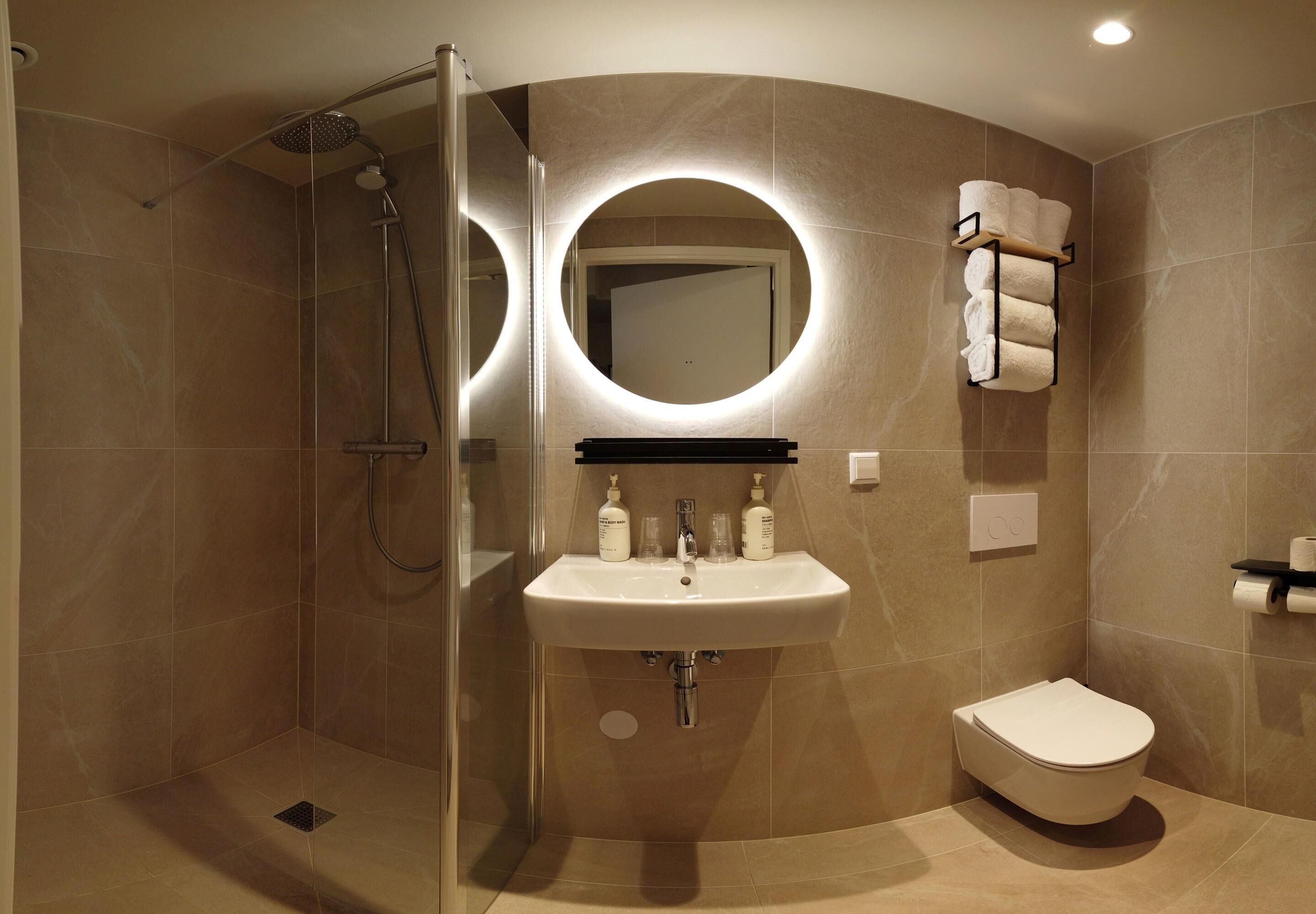 A modern bathroom with a glass-enclosed shower, a round mirror, a sink with two bottles, and a toilet.