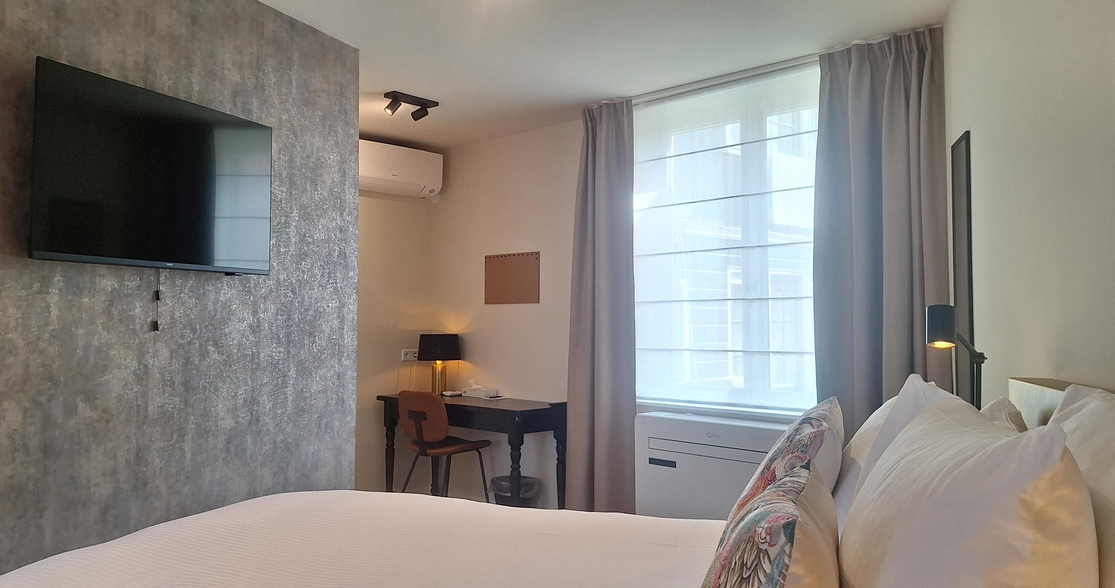Une chambre d’hôtel comprenant un lit, un bureau avec une chaise, une télévision, un appareil de climatisation et une fenêtre avec des rideaux.