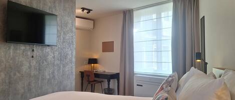 A hotel room with a bed, a desk with a chair, a TV, an air conditioning unit, and a window with curtains.
