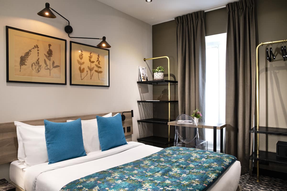 A neatly arranged hotel room with a bed, a desk, and a chair. There are framed botanical prints on the wall, a small plant on the desk, and a shoe rack by the window.