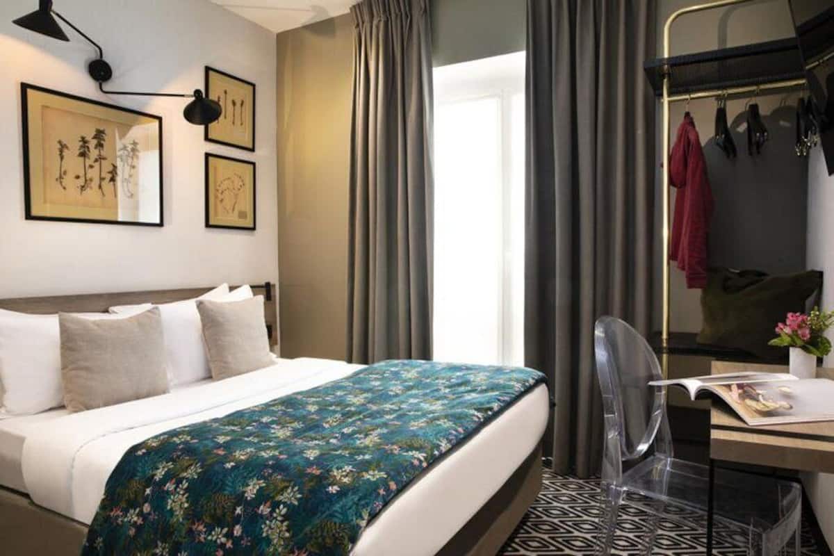 A hotel room with a bed, a desk with a chair, a mirror, and artwork on the wall.