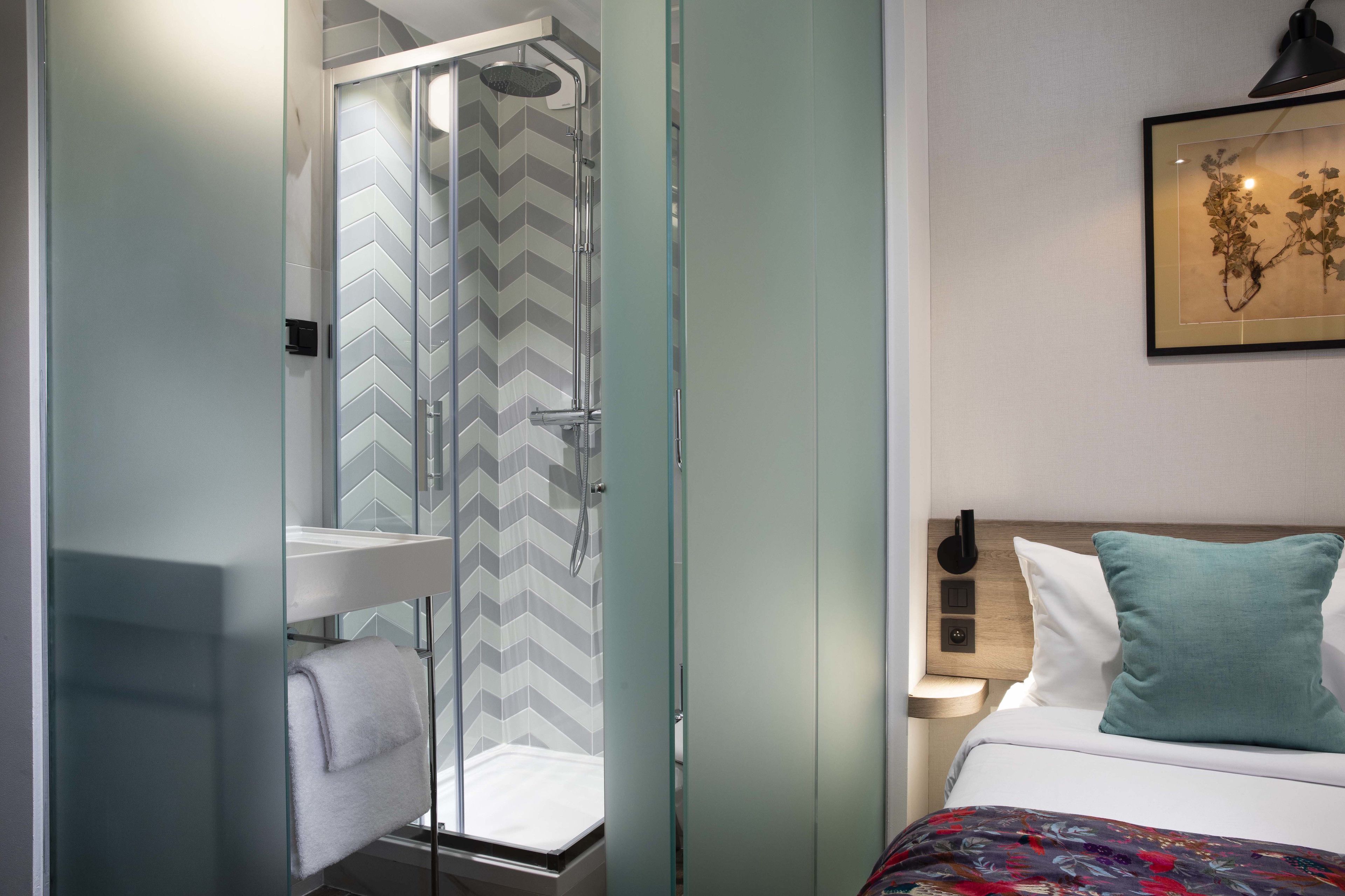A hotel room with a bed, a nightstand, a wall-mounted lamp, a framed artwork, and a shower with a glass door.