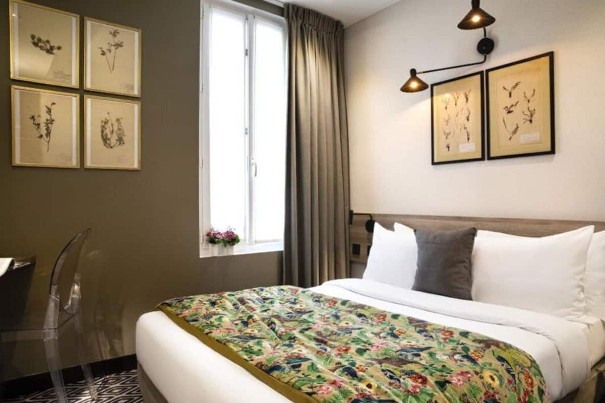 A hotel room with a bed, a desk, and a window with curtains. There are framed botanical prints on the wall.