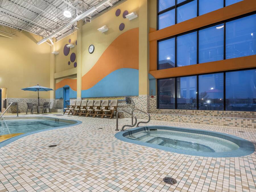 An indoor swimming pool with tiled floors, a clock, and a seating area with chairs and an umbrella.