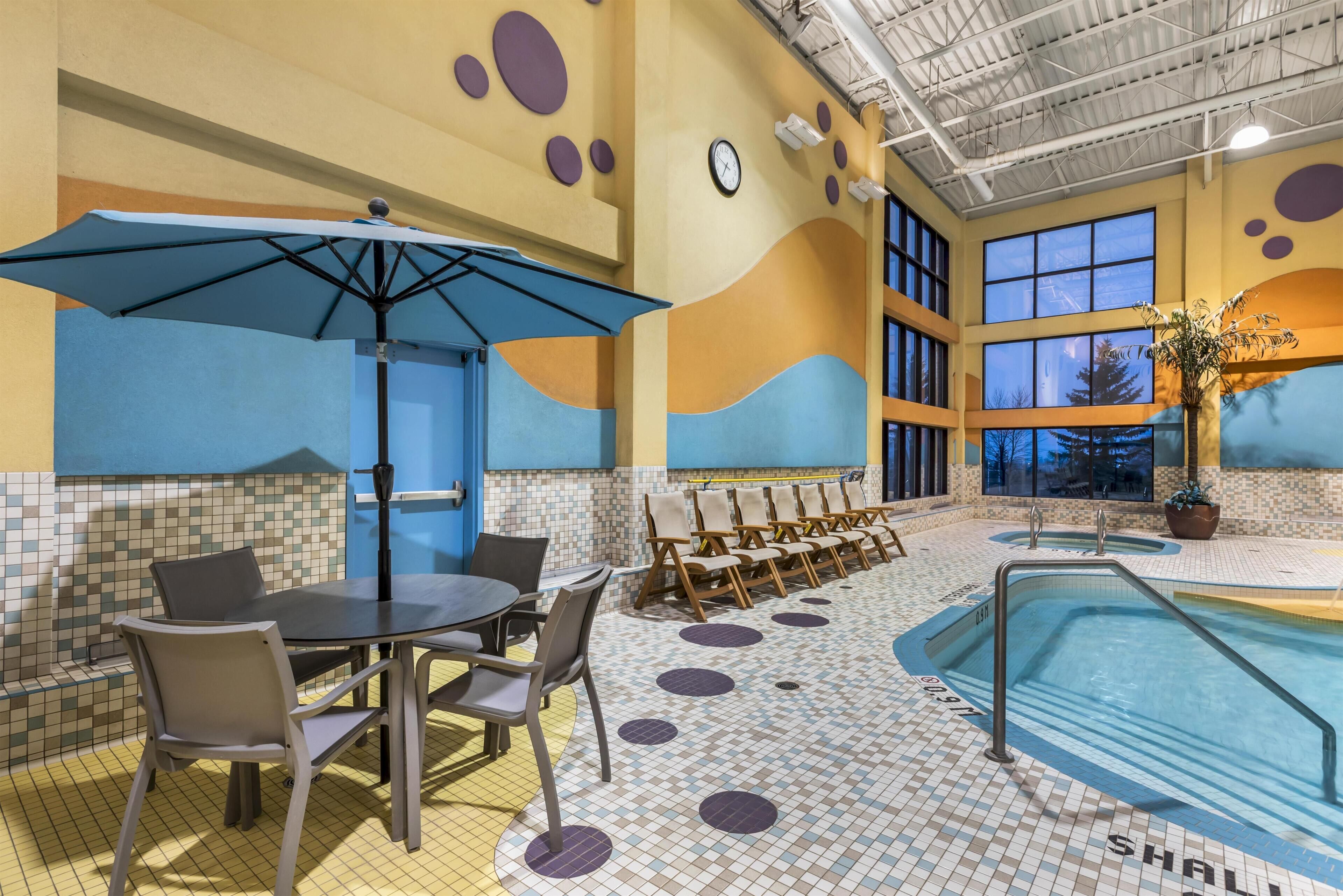 An indoor pool area with a dining table and chairs, a pool with a safety railing, and a clock on the wall.