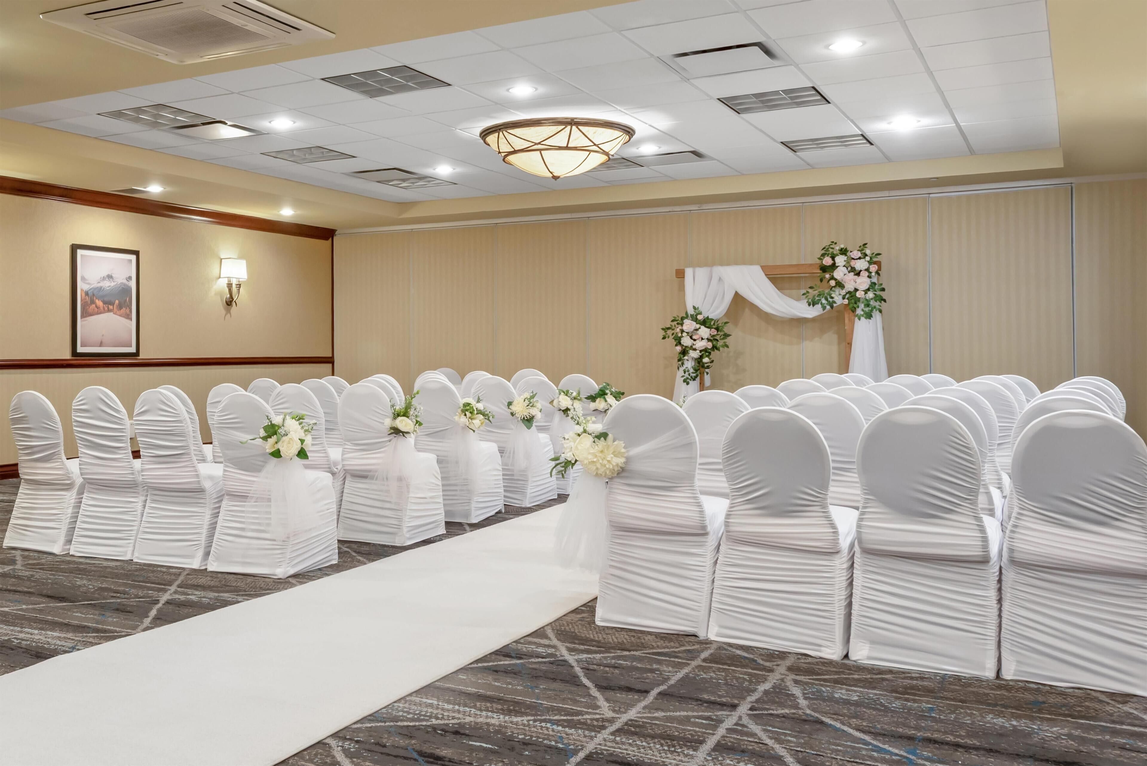 Un salón de conferencias con filas de sillas blancas, un pasillo central blanco y una decoración floral en la pared.