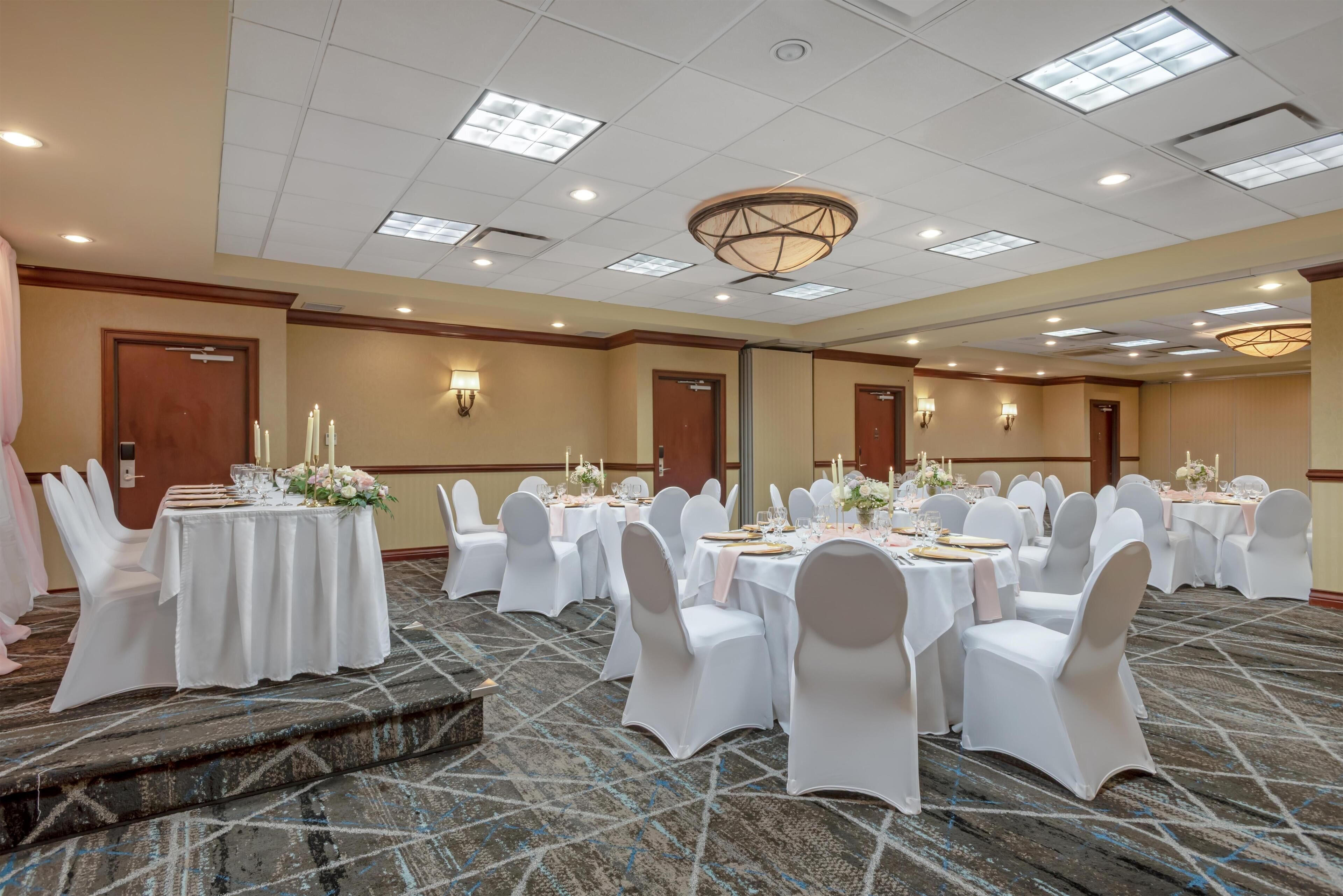 Un salón de banquetes con mesas redondas, sillas blancas y piso alfombrado.