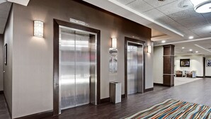 A modern hotel lobby with two elevators, a patterned rug, and a reception area with chairs and a small table. - Best Western Plus Port O'Call Hotel (Calgary)