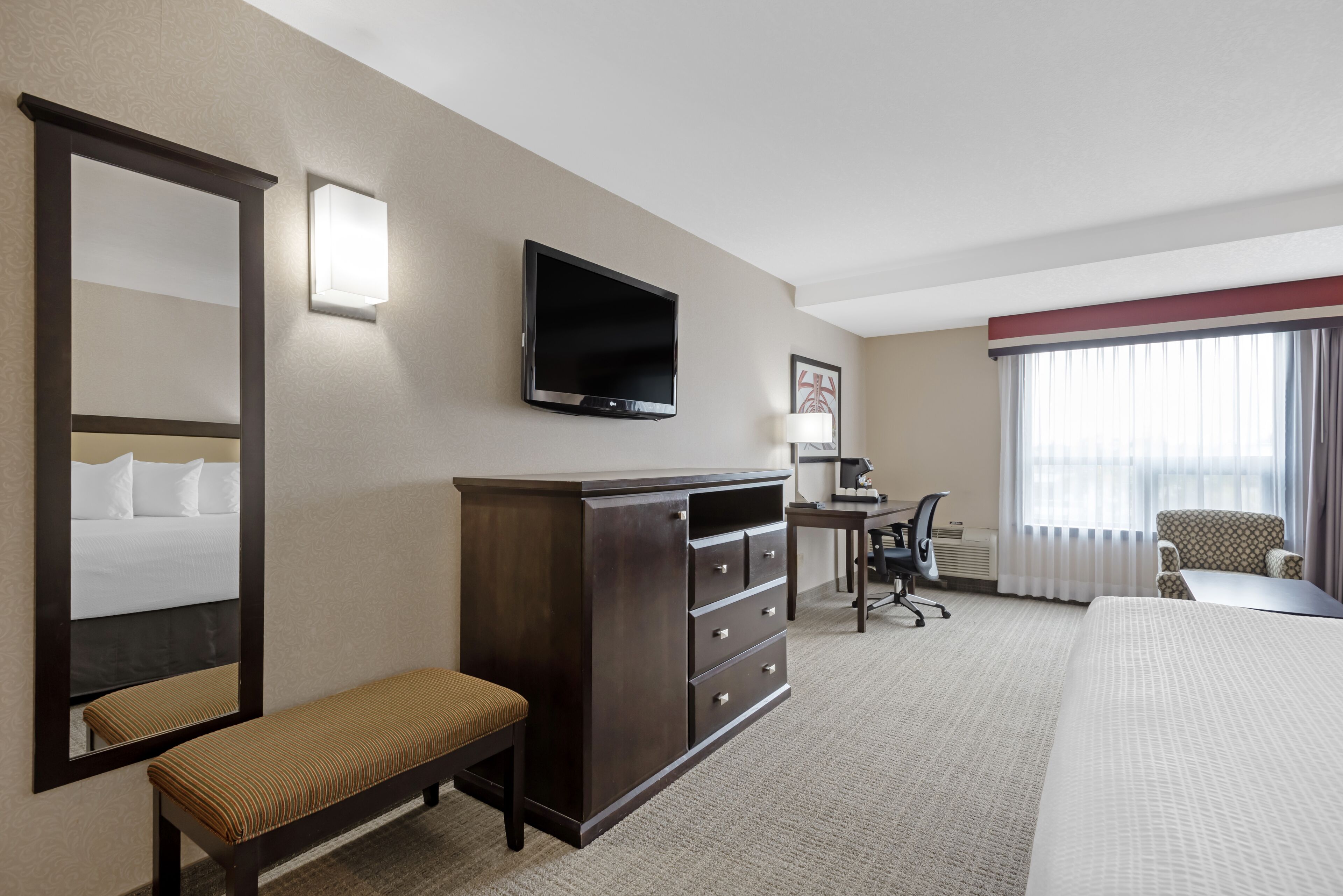 A hotel room with a large bed, a desk with a chair, a television, a mirror, and a window with curtains.