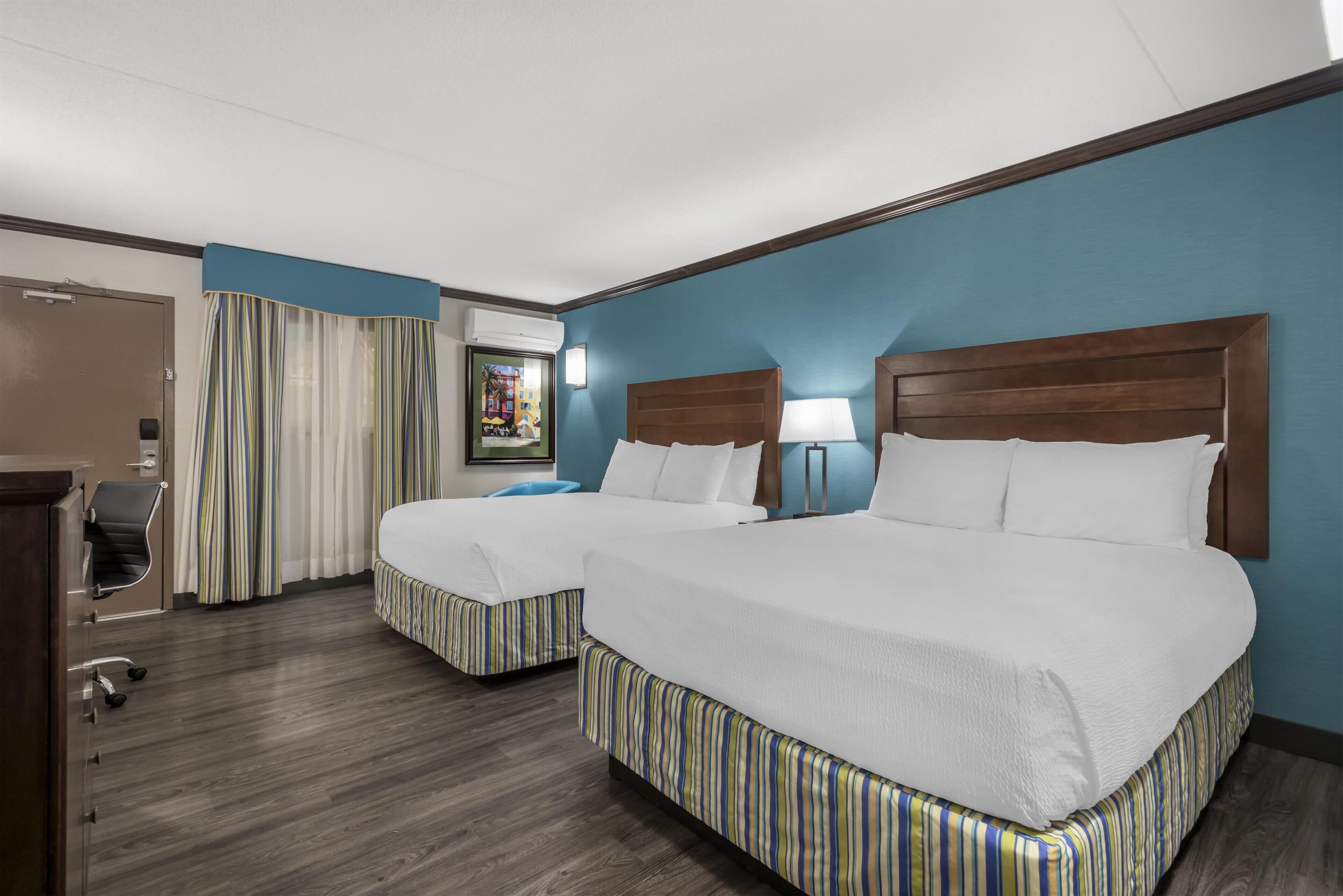 Una habitación de hotel con dos camas, un escritorio, una silla, una lámpara y un cuadro en la pared.