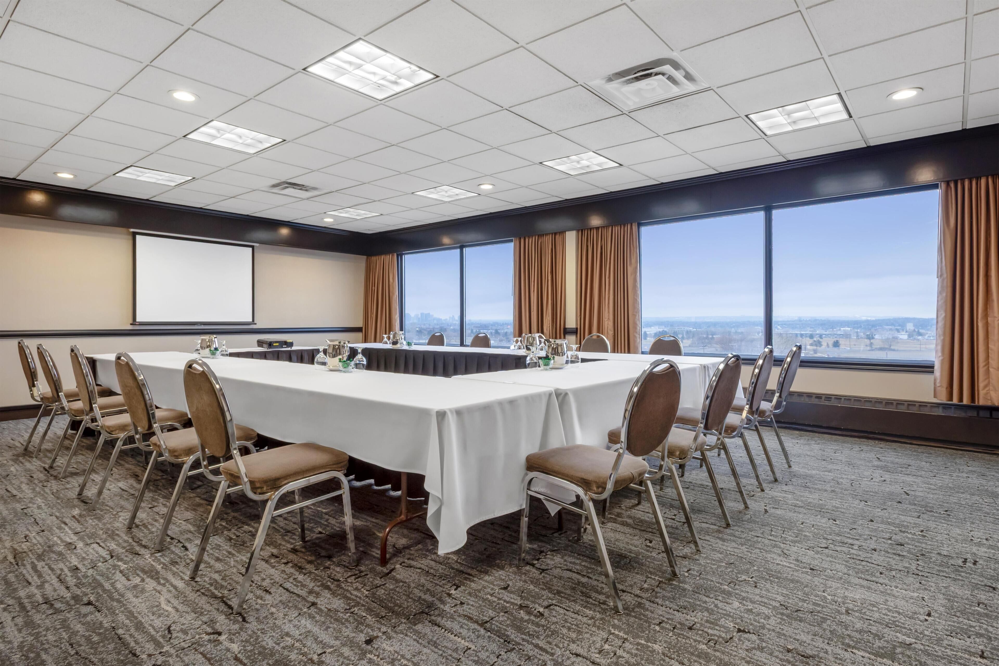 A conference room with a long table, chairs, a projector screen, and large windows with a city view.