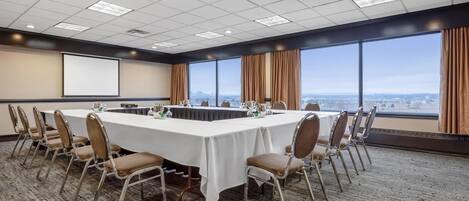 A conference room with a long table, chairs, a projector screen, and large windows with a city view.