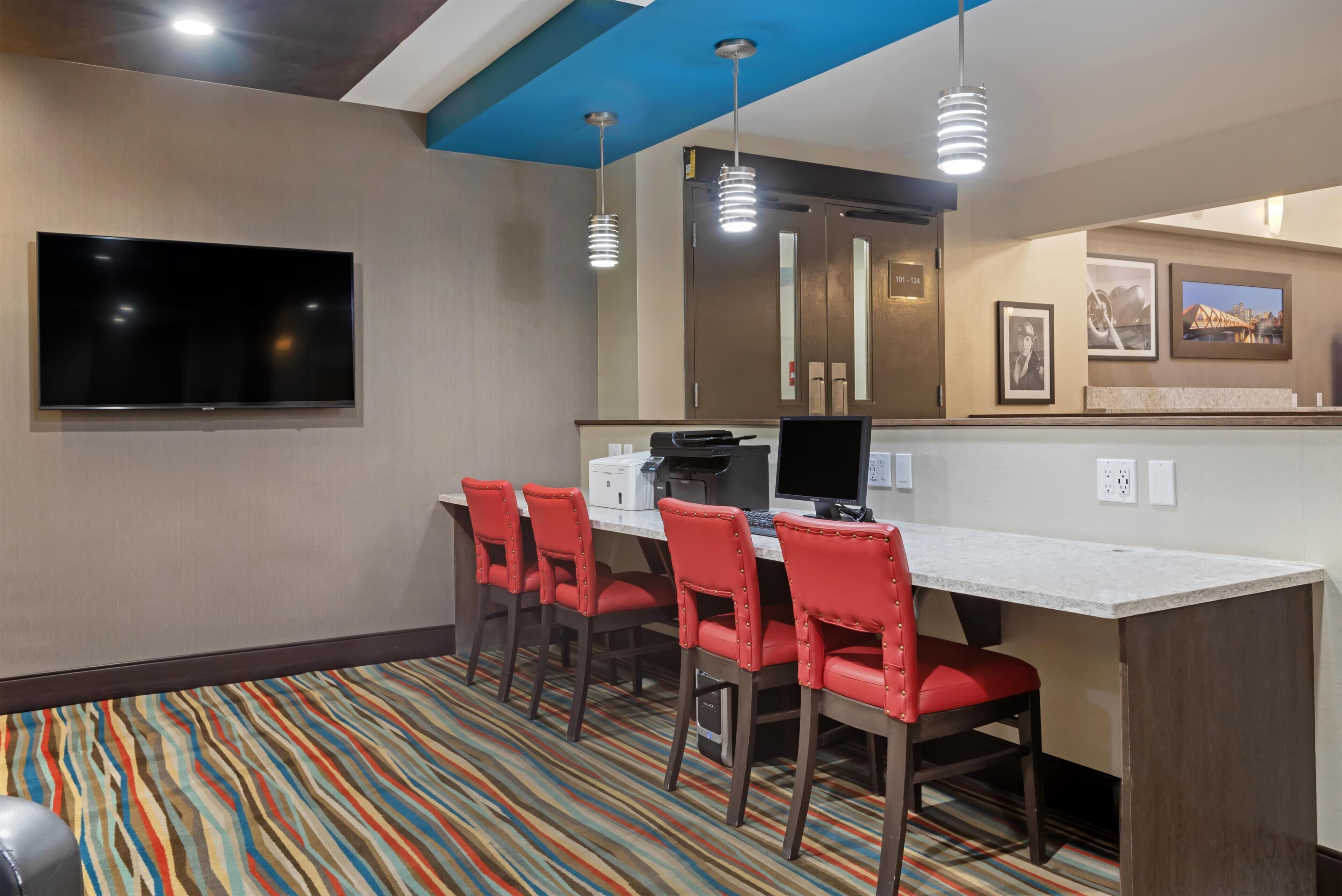 A hotel room with a flat-screen TV, a desk with a computer, four red chairs, a patterned carpet, and a blue ceiling.