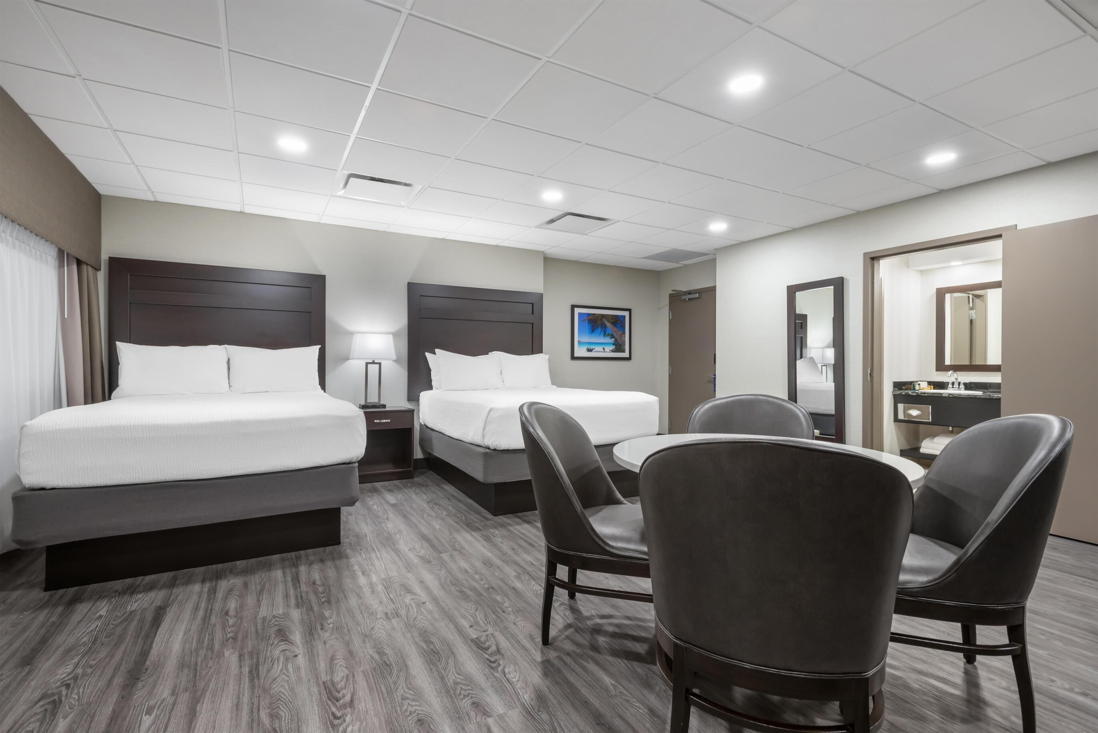 A hotel room with two beds, a desk, chairs, a TV, and a bathroom with a mirror.