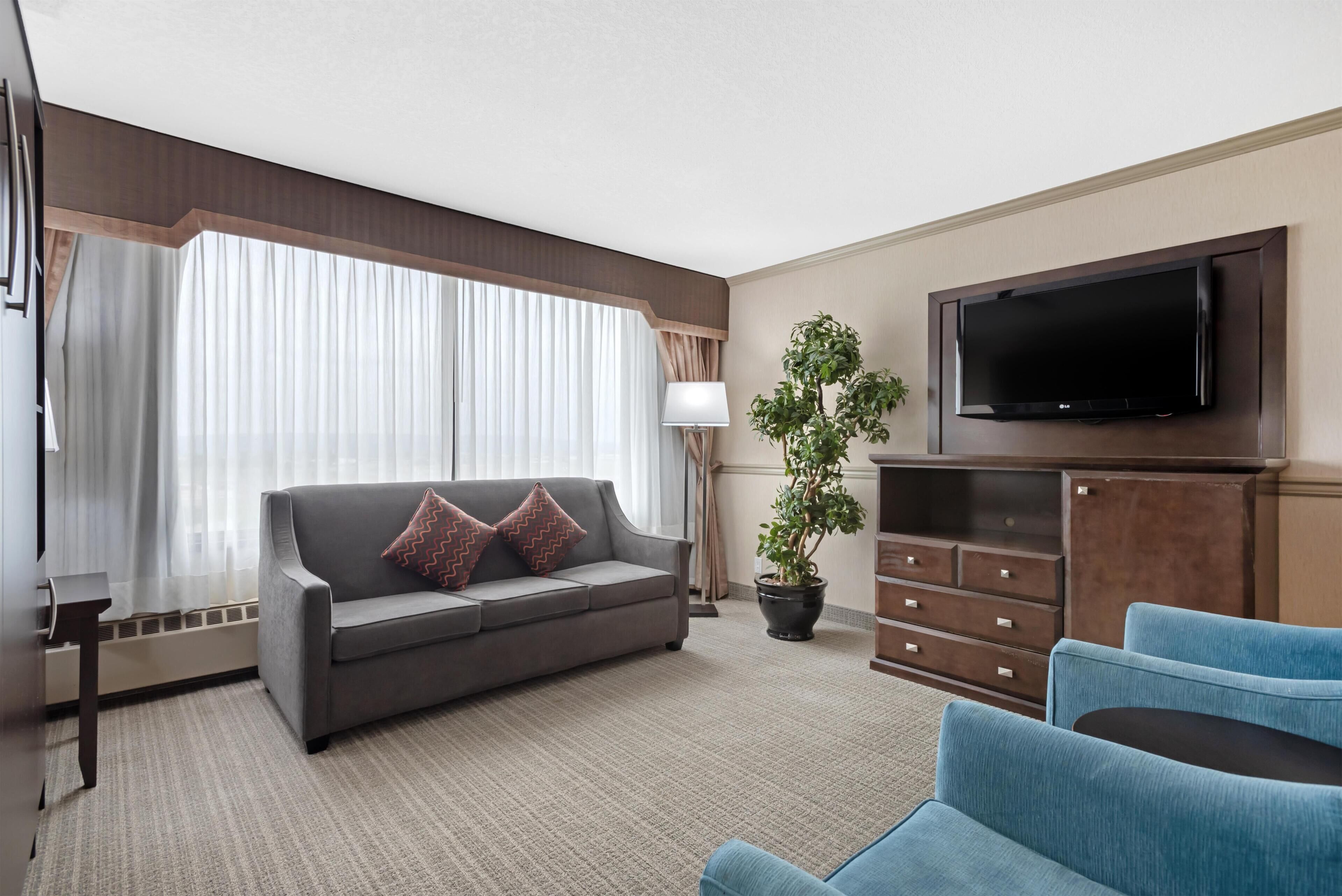 A hotel room with a grey sofa, blue armchairs, a flat-screen TV, a potted plant, and a large window with curtains.