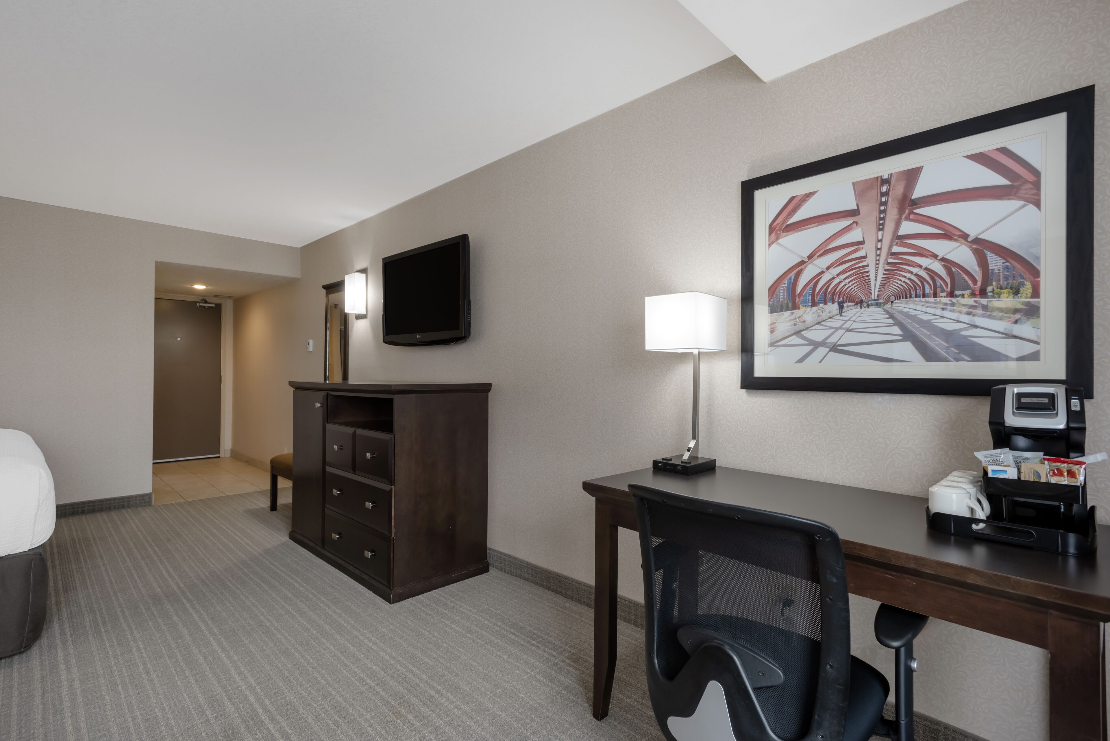 A hotel room with a desk, chair, TV, lamp, and a large framed picture on the wall.