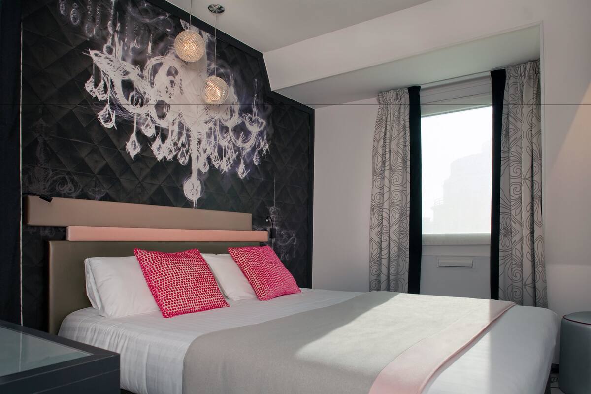 A modern hotel room with a large bed, a decorative wall, a chandelier, and a window with curtains.