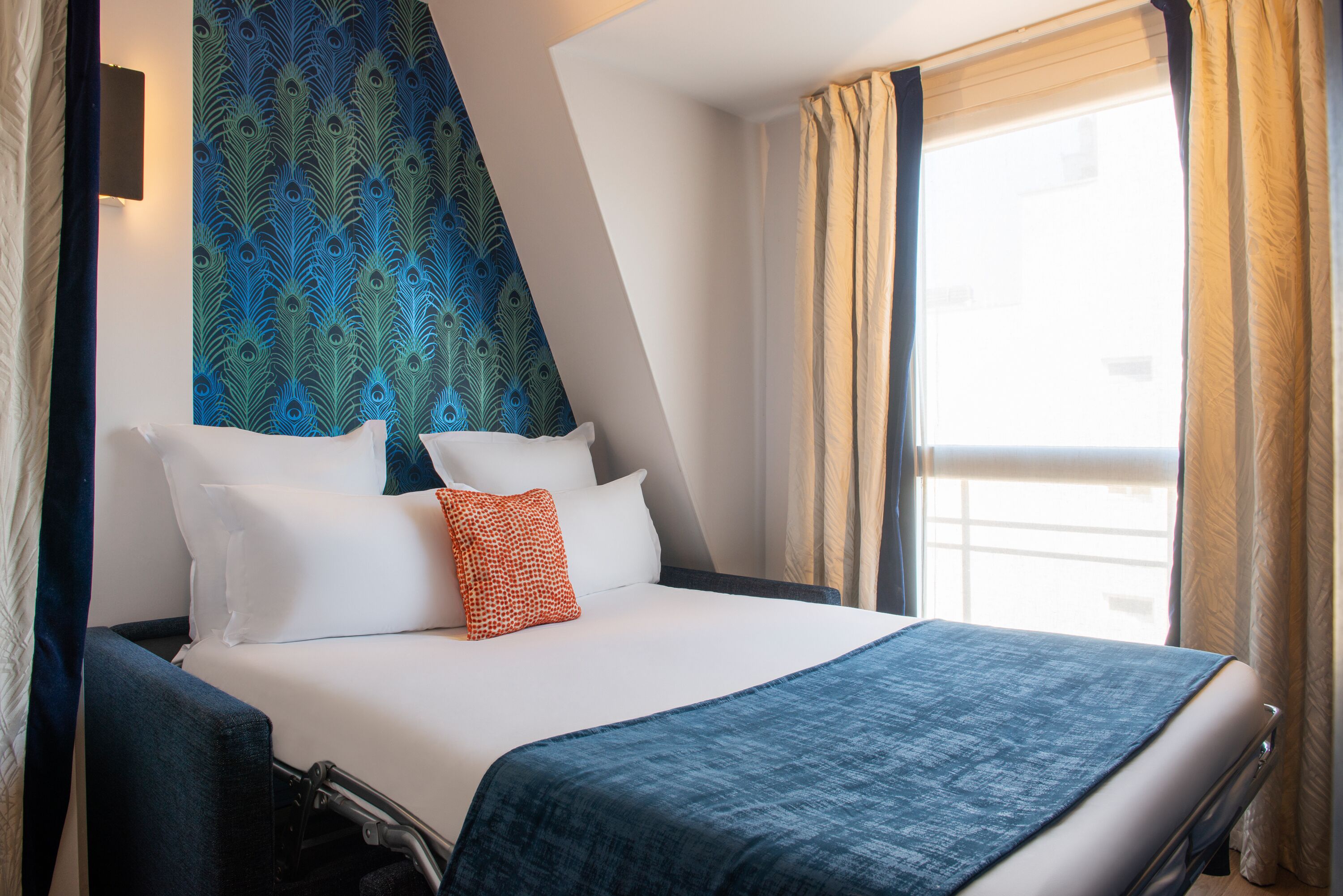 A hotel room with a bed, pillows, a blanket, a headboard with a peacock feather pattern, and a window with curtains.
