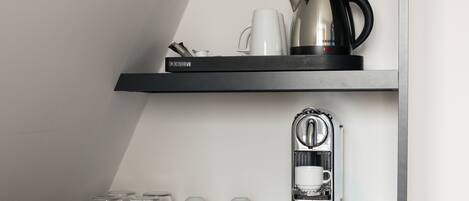 Un área de cocina con cafetera, hervidor y repisa con tazas y una taza.