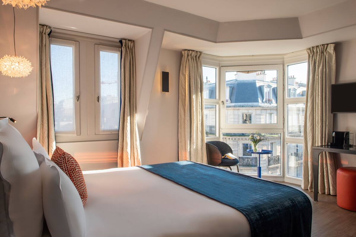 A hotel room with a large bed, a desk with a chair, a television, and a view of the city through large windows.
