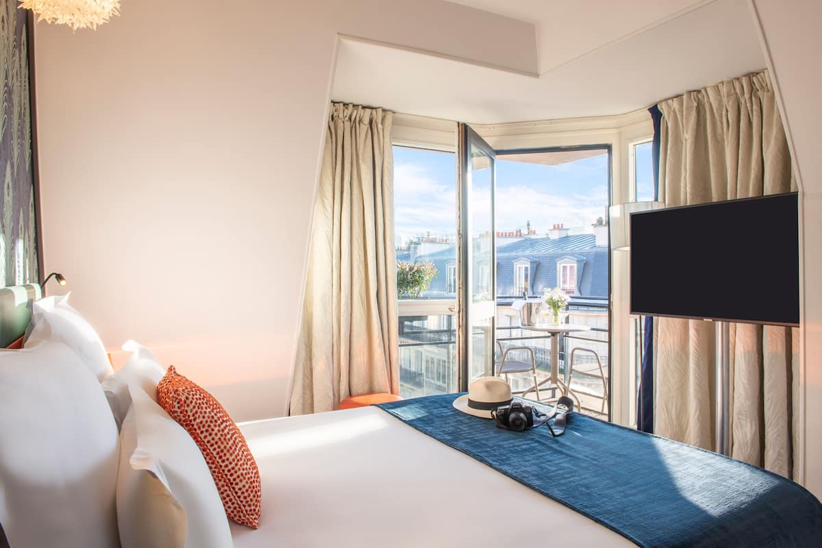 A hotel room with a large bed, a flat-screen TV, a view of the city through a large window, and a balcony with a table and chairs.