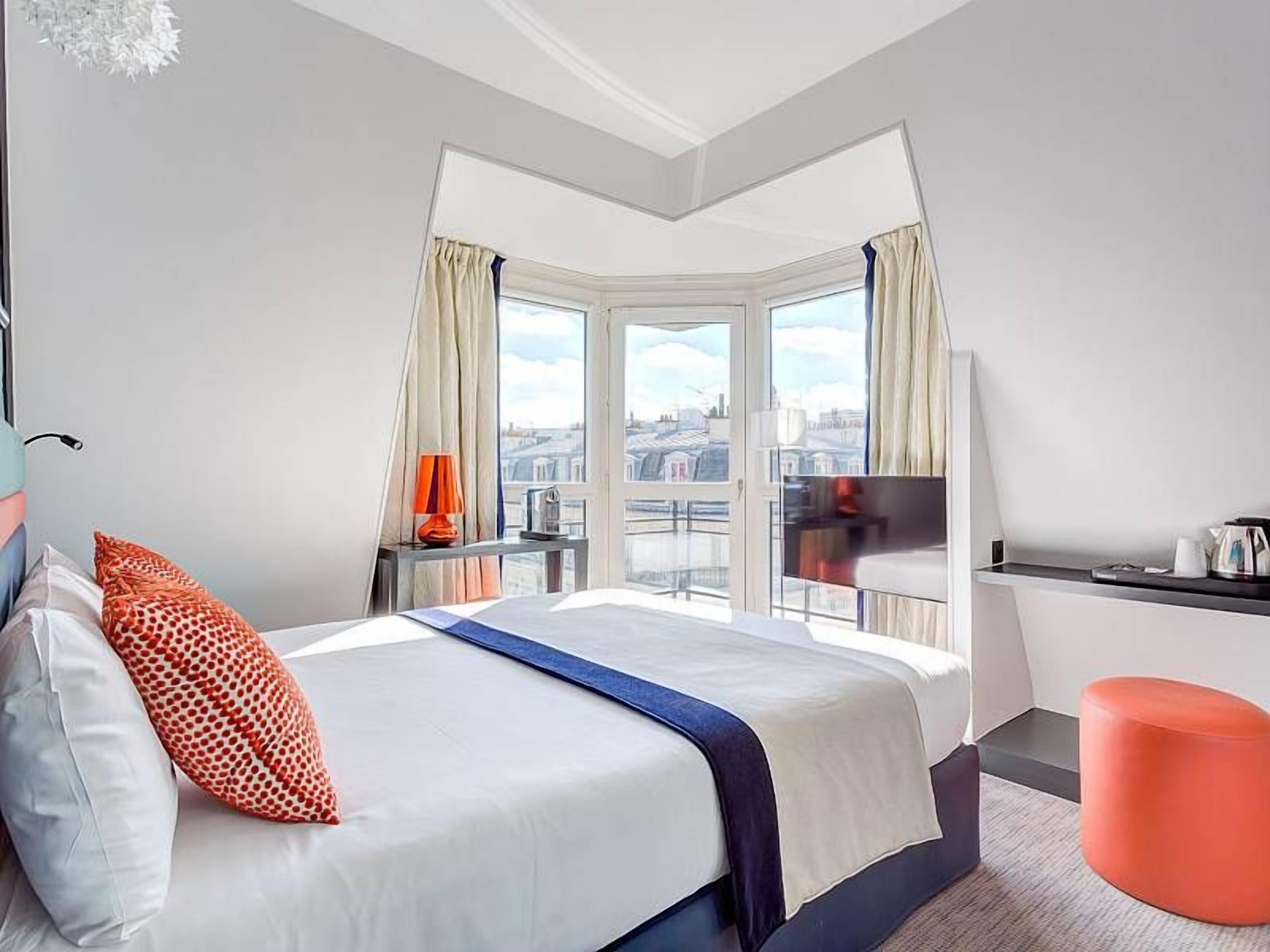 Une chambre d’hôtel avec un grand lit, une vue sur la ville, une petite table avec une lampe et un pouf rouge.