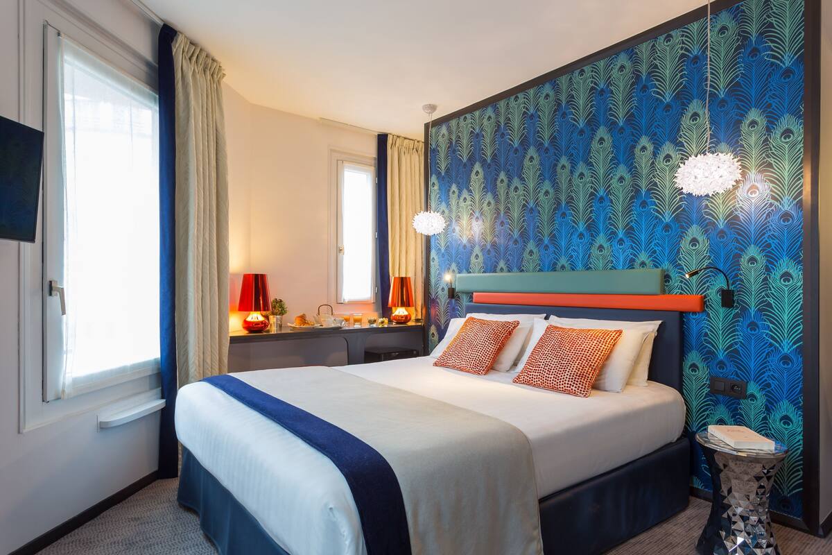 A hotel room with a large bed, a desk with a lamp, a peacock-patterned wall, and a small side table.