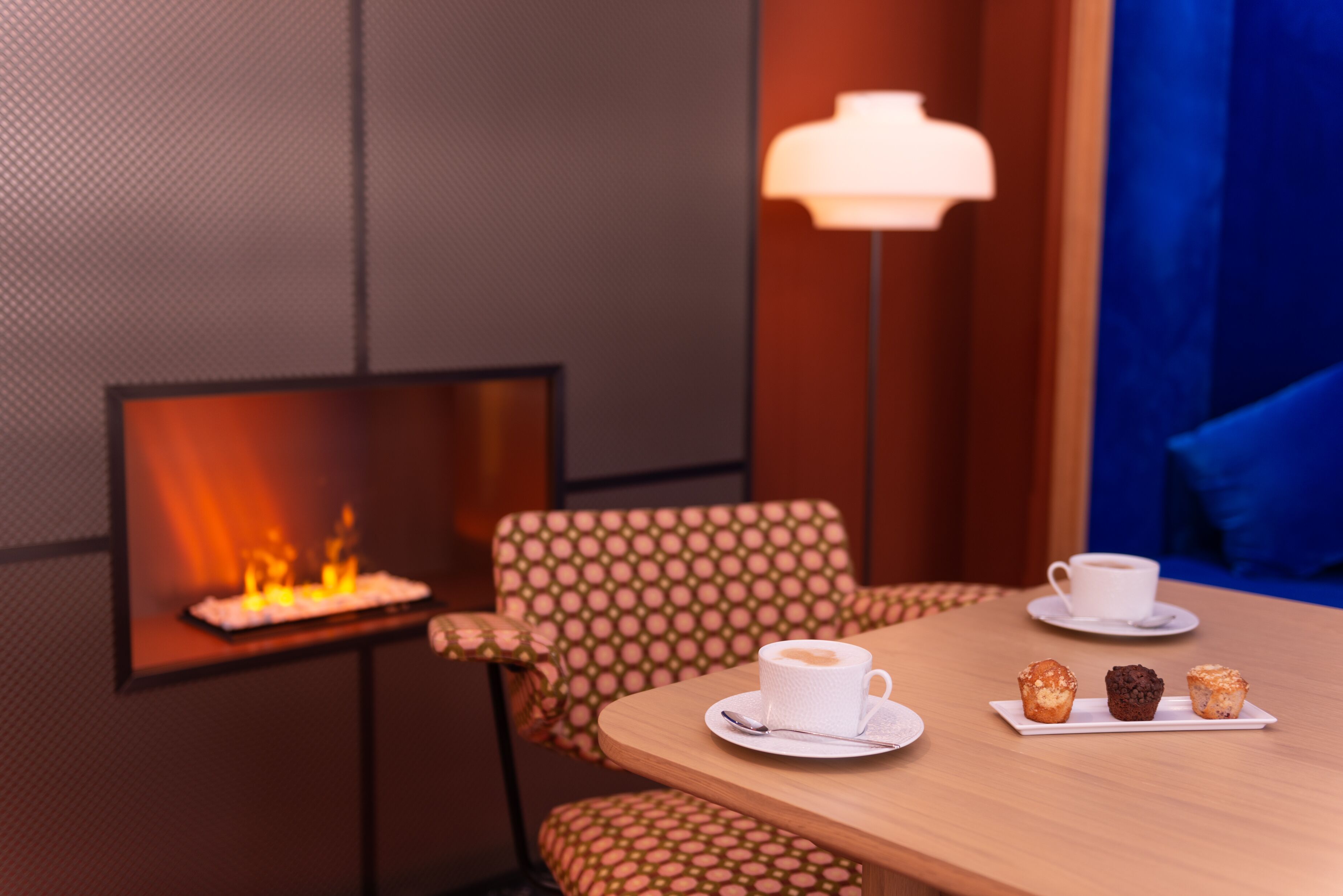 A cozy room with a fireplace, a table with a cup of coffee and pastries, patterned chairs, and a lamp.