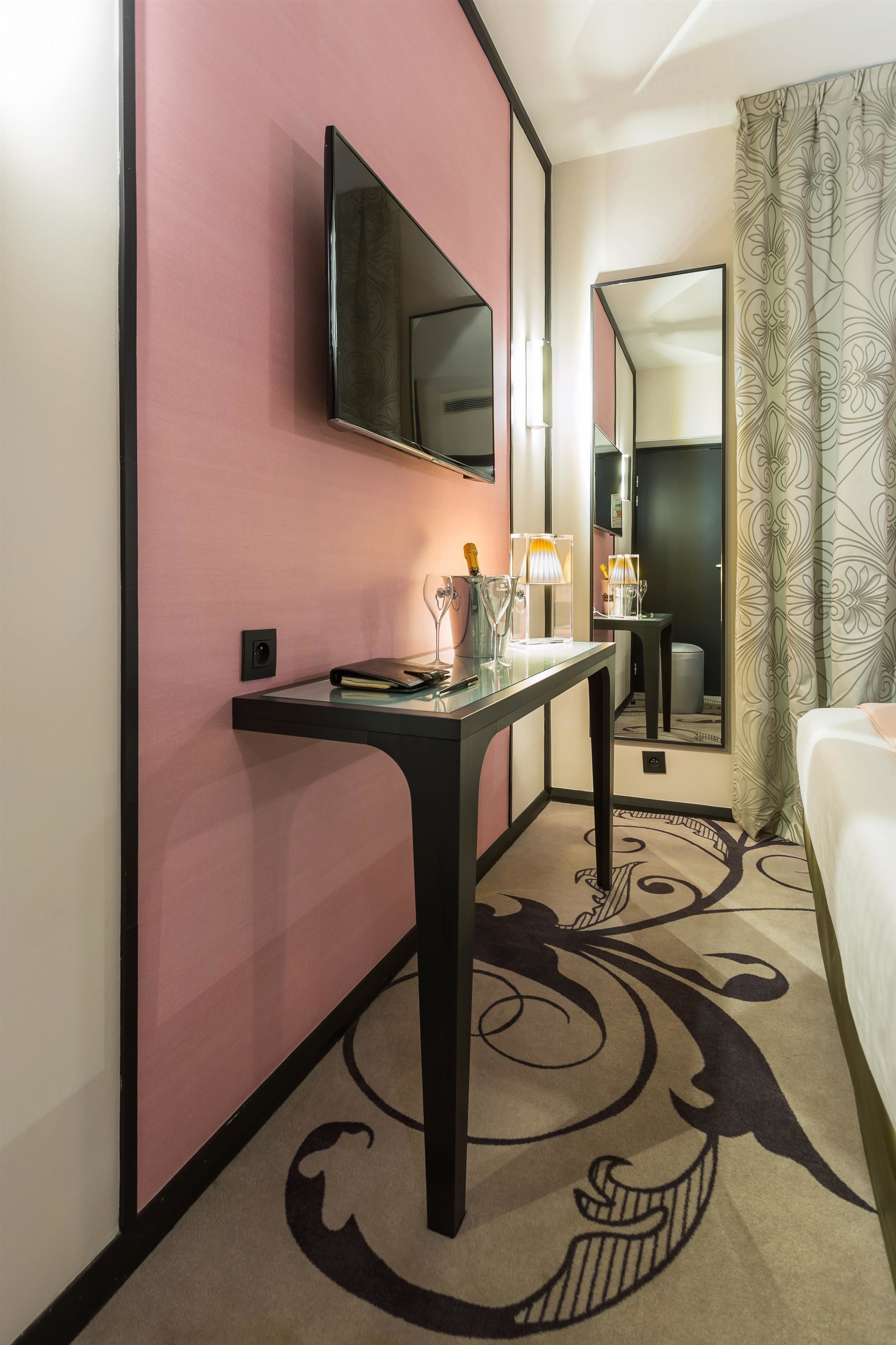 A hotel room with a pink accent wall, a flat-screen TV mounted above a desk, a bed with a patterned bedspread, and a large mirror reflecting the room.