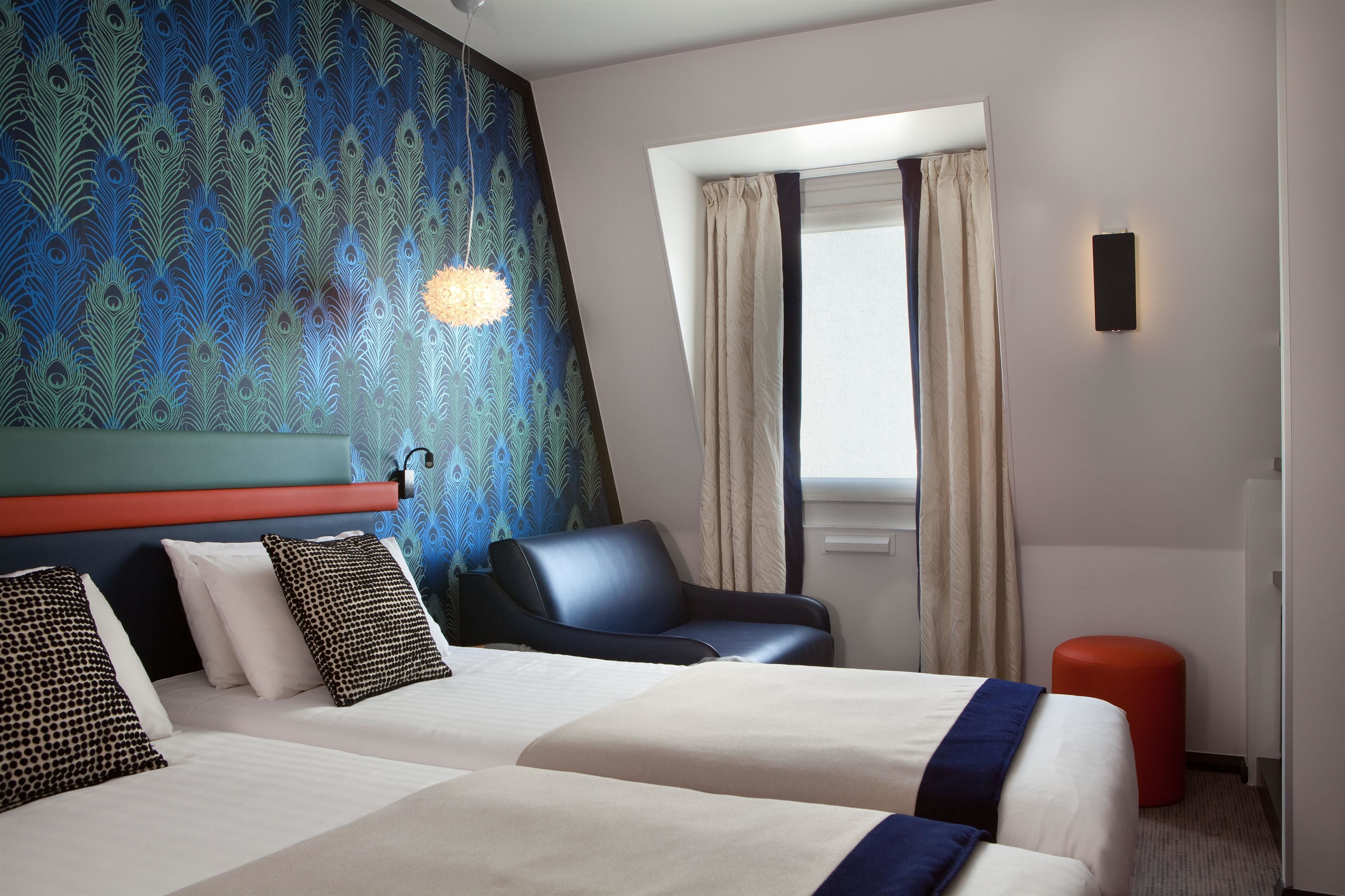 Una habitación de hotel con una cama, una silla, un pouf rojo y una ventana con cortinas.