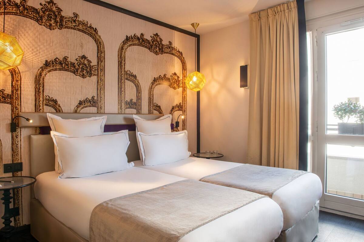 A hotel room with a large bed, two bedside tables, ornate wall mirrors, and a view through a window with curtains.