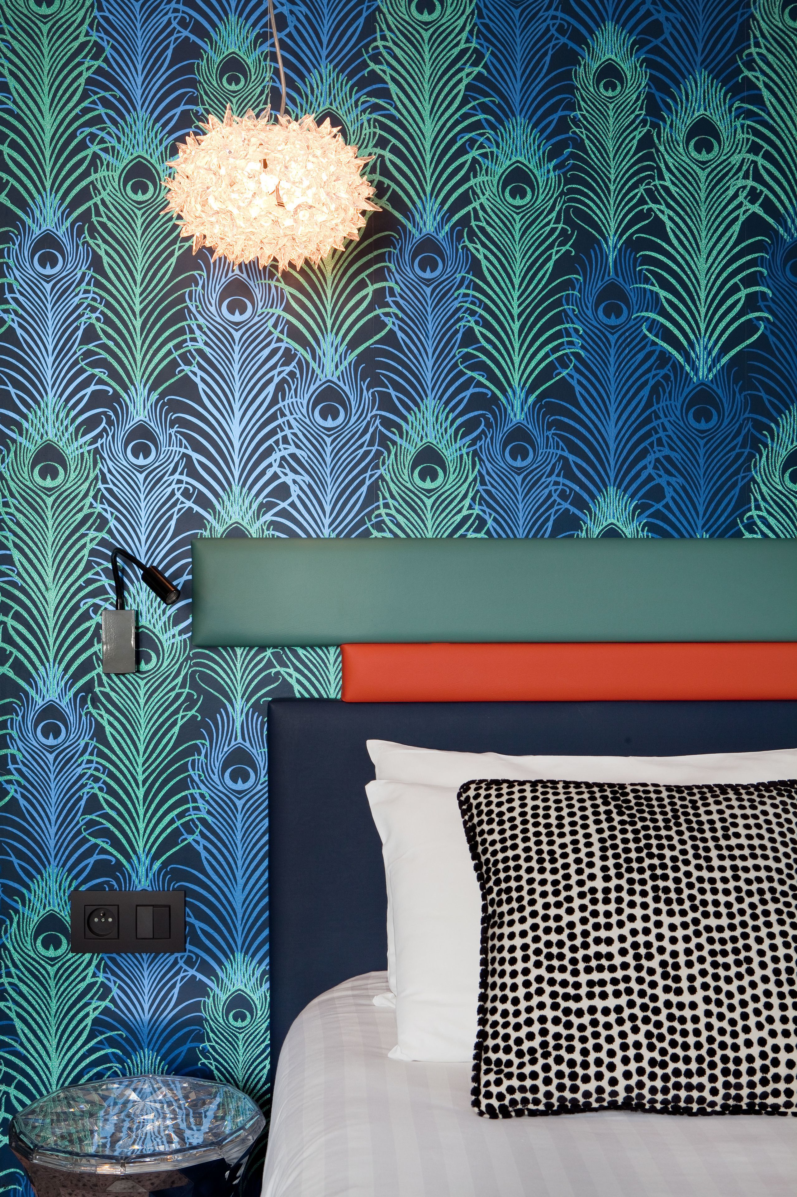 Un dormitorio con papel tapiz estampado con plumas de pavo real, una lámpara redonda, una cama con almohadas blancas y negras y una mesita de noche con superficie de vidrio.