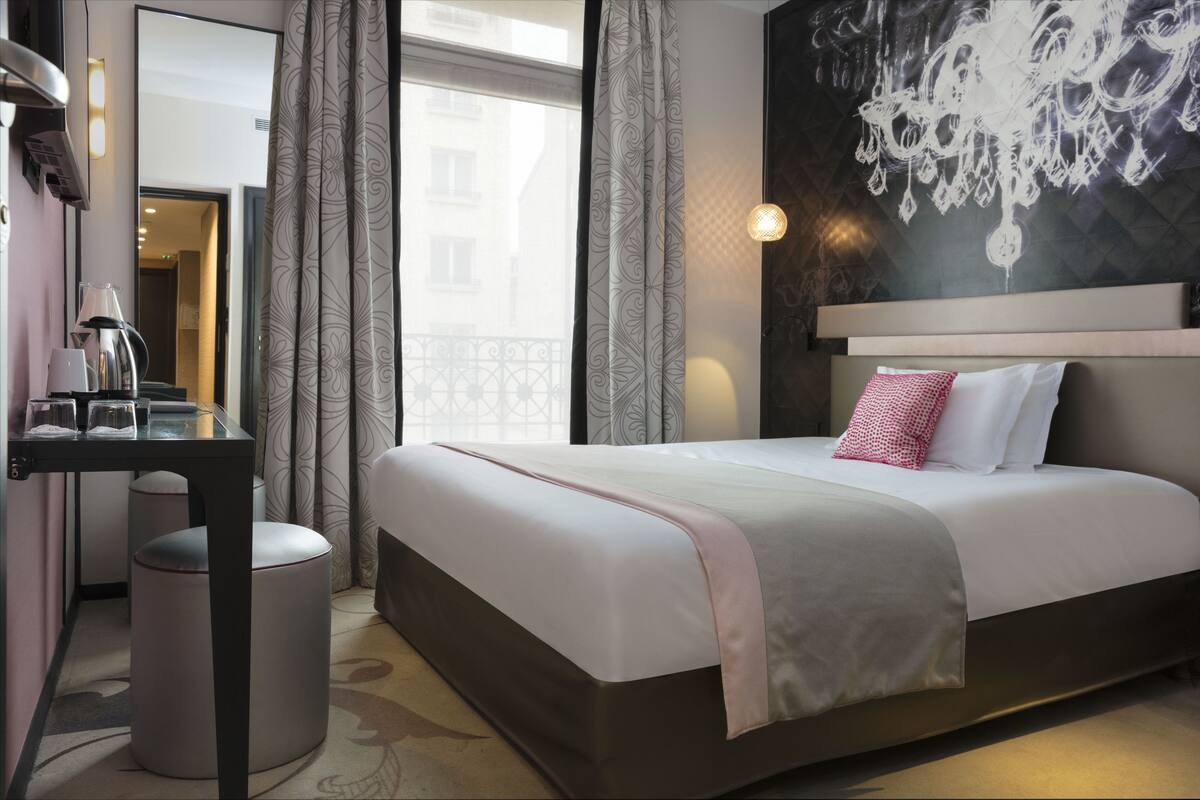 A modern hotel room with a large bed, a desk with a lamp, and a view of the city through the window.