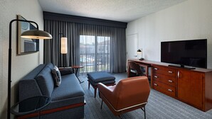 Une chambre d’hôtel équipée d’un canapé, d’un pouf, d’un bureau en bois, d’une télévision et d’une grande fenêtre avec des rideaux.